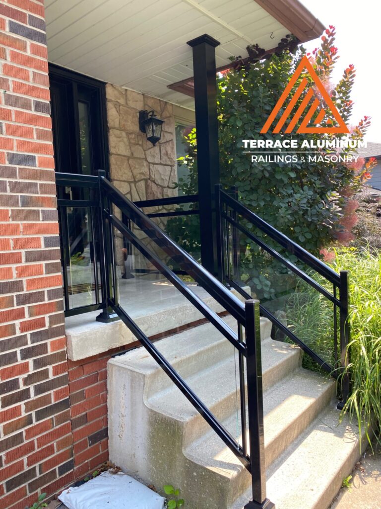 Black aluminum and glass railing installed on a front porch in Oakville by Terrace Aluminum Railings & Masonry. Clear tempered glass panels with black aluminum framing on concrete steps for a modern exterior look.